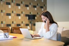 Focused female designer discussing project with customer on phone, sitting at table with laptop and blueprints and typing. Medium shot. Multitasking concept